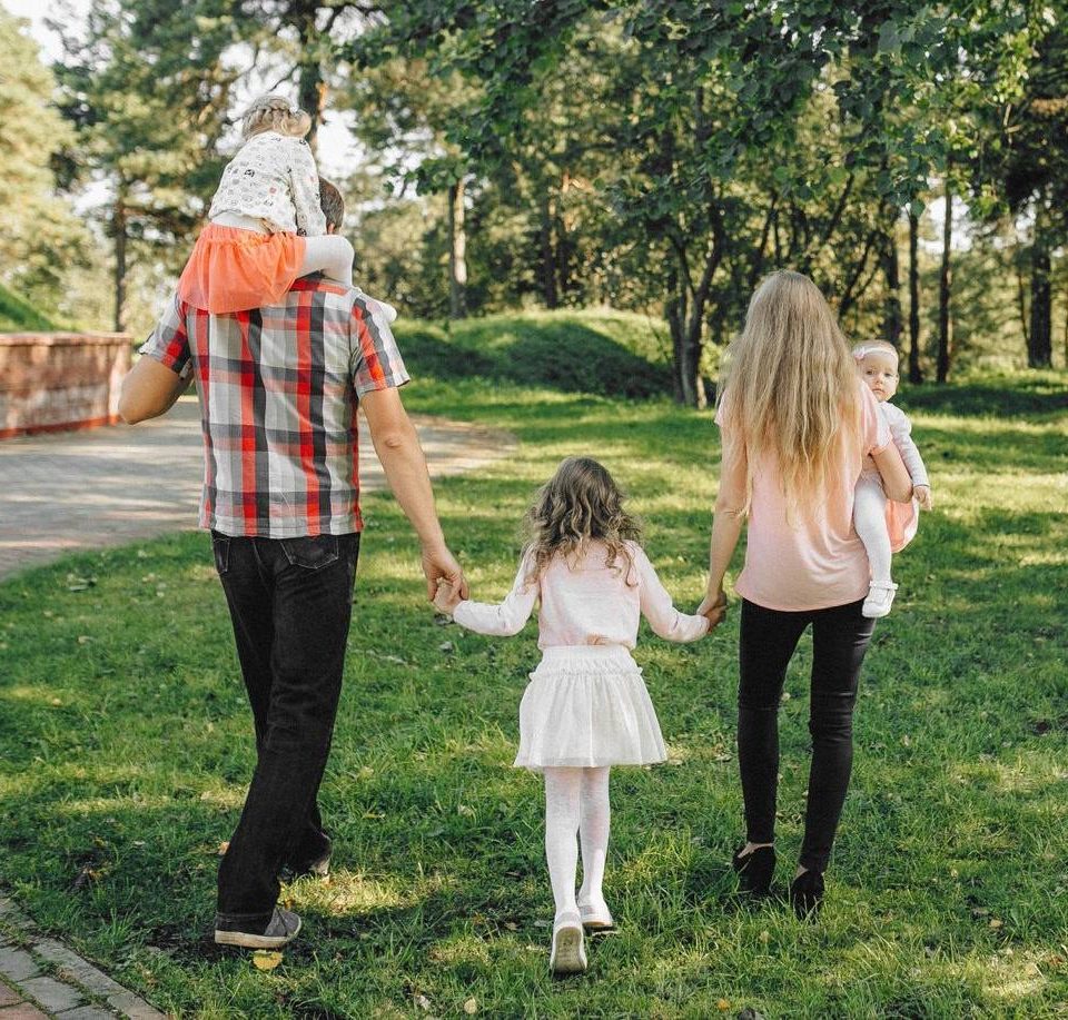 Família de costas caminhando em um parque: pai com uma criança nos ombros, mãe com o bebê no colo e uma menina de mãos dadas